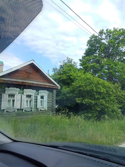 This media file captures a view from inside a vehicle, looking out at a traditional wooden house in Staryi Pokrov, Russia. The house, with its green wooden walls and ornate white window shutters and trim, appears to be quite old. The roof is made of dark wood, and a small window is visible on the gable.
The house is surrounded by tall, green grass and lush trees, suggesting a rural or suburban setting in a temperate climate. The sky is partly cloudy, with patches of blue visible, indicating daytime and fair weather. A power line runs diagonally across the upper portion of the frame.
The perspective suggests the viewer is looking out through a windshield, with a portion of the car's dashboard visible in the foreground. There are no people visible in the image, and the overall impression is one of a quiet, picturesque rural landscape. The time of day appears to be late morning or early afternoon. No text is visible in the image.