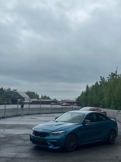 In the city of Kovalyovo, Russia, an overcast sky looms over an outdoor setting, possibly a race track or industrial area. In the foreground, a teal BMW M2 is parked on a wet asphalt surface. The car is positioned at an angle, with its front and driver's side visible. To the left of the BMW, a person stands near a metal fence, facing away from the camera. Behind the person and the fence, a large, grey industrial building with a curved roof is visible. Several shipping containers are stacked behind the fence, and more vehicles, including red cars and a white truck, are present in the background. Dense green trees line the right side of the scene.