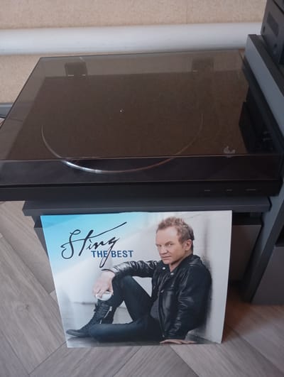 The image shows a vinyl record album cover featuring the artist Sting, titled "The Best," placed in front of a turntable. Sting is depicted wearing a black leather jacket and dark pants, holding a white cup. The album cover is positioned on a wooden floor. The turntable is a dark-colored electronic device with a clear lid. The overall scene appears to be an indoor setting, likely a room in a residential or studio environment. There is no information within the image that indicates the location is in the city of Vysokaya Gora, Russia.