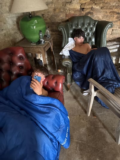 Two people are sleeping on leather armchairs. One person is lying on a green armchair with a blue sleeping bag partially covering them and a white towel under their head. The other person is lying on a red armchair completely covered with a blue sleeping bag while holding a blue smartphone. A lamp is next to the red armchair. The floor appears to be concrete.