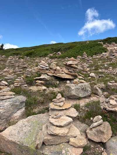 The image depicts a mountainous landscape under a bright blue sky with scattered white clouds. The foreground and midground are covered in numerous cairns, which are stacks of stones. These cairns vary in size and height, with some appearing as simple piles and others as carefully balanced structures. The stones used for the cairns are predominantly light brown and tan, with some exhibiting rough textures and lichen growth. Patches of green grass and low-lying vegetation are interspersed between the stones. In the background, a grassy slope rises, covered in dense green bushes. A single evergreen tree is visible on the far left, near the base of the slope. The scene appears to be a hiking trail or a natural landmark, possibly in a high-altitude area. No people are present in the image. The location is identified as Urtijëi - St. Ulrich in Gröden - Ortisei, Italy, suggesting a region known for its mountainous terrain and outdoor activities.