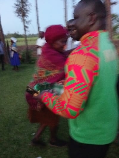 In a somewhat blurry outdoor shot, a man in a bright green shirt with patterned sleeves, embracing an older woman with a red headscarf. She wears a patterned shawl and brown skirt. Several other blurred people are visible in the background, one in a white shirt. A woman in a blue dress and white head covering is further back. The ground is green with trees in the background.