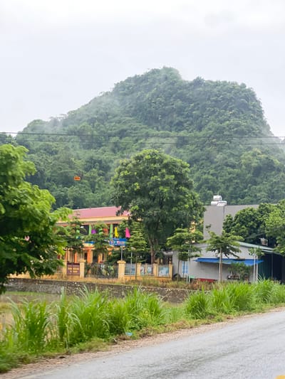 This image captures a scene in Tân Thanh Commune, Vietnam, featuring a lush green, mist-covered mountain dominating the background. In the mid-ground, a school building with a reddish-orange roof is visible, adorned with colorful flags and what appears to be a banner with Vietnamese text. In front of the school, a yellow fence encloses the property. To the right of the school, a modern, light-colored building with a blue awning stands, and further to the right, another structure with a dark roof is partially visible. A body of water, likely a canal or river, runs along the front of the school, bordered by a stone wall and dense, tall green grass. A paved road, wet from rain, occupies the foreground, with more green grass and vegetation lining its edge. The sky is overcast and hazy, contributing to the muted tones of the landscape.