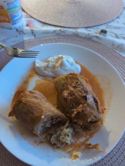 This is a top-down, eye-level medium shot of a white plate filled with two cabbage rolls in a tomato-based sauce, accompanied by a dollop of sour cream. A fork rests on the lower left side of the plate, positioned to serve the food. The cabbage rolls appear to be filled with a mixture of ground meat and rice. The sauce is a vibrant orange-red, and some of it has spread around the rolls and onto the plate. The sour cream is a bright white, contrasting with the sauce.

The plate is set on a light-colored tablecloth with a floral pattern. To the left of the plate, a textured, circular placemat is partially visible. A cylindrical object with visible text is also in the lower left corner of the frame. To the right of the plate, a woven placemat with a diamond pattern is visible.

The lighting suggests an indoor setting, possibly daytime, with natural light contributing to the illumination. The overall scene depicts a meal being served or eaten. No people are visible in the image.

Visible text includes "Słupsk, Poland" on a label of what appears to be a food product in the lower left corner. Additionally, partial text reading "8%" is visible on the same product label.