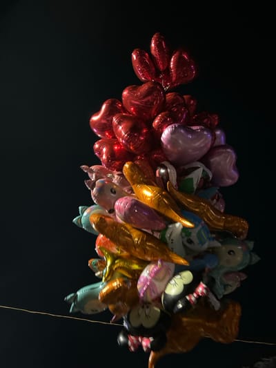 A cluster of brightly colored balloons is hanging from a string in the night sky, photographed from below. There are red, pink, blue, gold, and white balloons. The location is near the Konak Square area of İzmir, Türkiye, based on the provided coordinates. The image is taken at night, and the background is a dark sky. The balloons are likely from a street vendor or celebration. There are no people or objects in the image besides the balloons and the string.