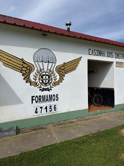An outdoor daytime scene depicts the front exterior of a single-story building in Praia do Ribatejo, Portugal. The structure features white walls with a dark green baseboard and a red corrugated metal roof, from which a black pipe topped with a silver cap protrudes. A small, dark-colored light fixture is mounted on the wall below the roofline.
On the left section of the white wall, a large painted emblem is visible: a grey parachute flanked by gold wings, encompassing a wreath that frames the Portuguese coat of arms. Below this emblem, the word "FORMAMOS" is painted in black capital letters. Directly beneath "FORMAMOS," the numbers "47156" are displayed on five individual white rectangular tiles with black borders.
To the right, an open, rectangular alcove is cut into the building's facade. Inside the alcove, a brown and black mountain bicycle, accented with red components on its seat and water bottle cage, is parked on a reddish-brown tiled floor. The interior walls of the alcove are dark. Above the alcove opening, the phrase "CASINHA DOS INSTRUTORES" is painted in black capital letters. A small white sign with black text, partially visible on the right interior wall of the alcove, reads "CENTRO DE INSTRUÇÃO AERO-TERRESTRE."
In the immediate foreground, a cracked concrete path runs parallel to the building. To the far left of the path, a grey plastic crate rests on the ground. Beyond the path, an area of green grass is visible. The sky overhead is clear blue with scattered white clouds. No individuals are present in the scene.