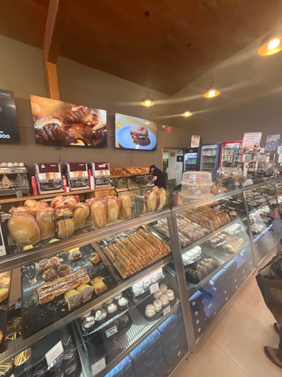 This image depicts the interior of a bakery or cafe located in Tolhuin, Argentina. The scene is dominated by a long array of illuminated glass display cases showcasing various baked goods and pastries.
The ceiling features light brown wooden planks with exposed beams and multiple black, industrial-style pendant lights emitting a warm yellow glow. The walls are painted a neutral grey-brown. Three large digital screens are mounted on the back wall. The left-most visible screen shows "UNION" and "AD 200." The central screen displays a close-up of glazed pastries or ribs in a dark sauce. The right-most screen features a hamburger on a blue plate against a yellow background.
Below the screens, a wooden counter holds a silver espresso machine on the left, followed by three red and silver "NESCAFE" branded hot beverage dispensers. Behind this counter, further back, are glass display cases containing loaves of bread, some individually wrapped. A male staff member, wearing a dark short-sleeved shirt, is visible behind a counter in the mid-ground, leaning over a display and appearing to arrange items.
In the background, two glass-fronted refrigerators, one with a blue frame and one white, contain various beverages. A white sign near the refrigerators displays "PROMO X KILO" and "€ 10,000," along with images of desserts.
The foreground is dominated by multi-tiered glass display cases. The upper shelf in the immediate foreground contains numerous light brown, cylindrical churros, likely sugar-dusted. Adjacent to this, on a black tray, are various pastries, including rectangular braided items with white icing and smaller individual cakes. The lower shelves within these cases feature an assortment of small, round white pastries, some with pink accents, darker cupcakes, and rows of light-colored pastries such as croissants and alfajores. Blue LED lighting illuminates the lower sections of these display cases. On the far right, the lower torso and arms of an individual, possibly a customer, wearing a dark jacket and holding a smartphone, are visible next to the display cases. The floor is covered with light-colored tiles.