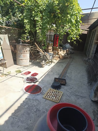 In the city of Aygezard, Armenia, an outdoor scene unfolds on a concrete surface under a canopy of grapevines. Several flat, round trays containing a reddish-brown substance, likely fruit pulp, are laid out to dry. To the right, a metal tray holds numerous small, circular, dried food items, possibly fruit slices or cookies. A black plastic tub rests on the ground nearby, and a larger red tub is partially visible in the foreground. In the background, a rusty metal barrel stands beside a wheelbarrow filled with gardening tools, suggesting agricultural activity. Stacked bricks and building materials are also present, along with a metal fence and what appears to be a punching bag. The overall environment is rustic and suggests food preparation or preservation is taking place.