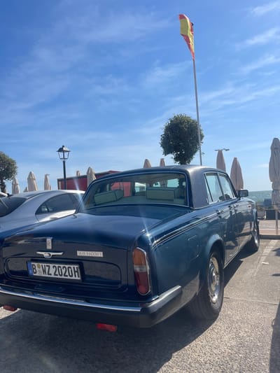 The image shows the rear of a dark blue Rolls-Royce Silver Shadow II parked near the harbor in El Puerto de Santa María, Spain. The Spanish flag is visible on a pole, and beach umbrellas are set up along the waterfront. The license plate reads "B WZ 2020H." The weather is sunny with a blue sky and light clouds.