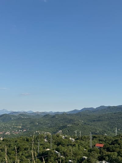 This is a landscape photograph taken in Dubovik, Montenegro. The image features a vast expanse of lush green hills and mountains under a clear blue sky. In the mid-ground, a small village with red-roofed houses is nestled among the trees. Several tall, silver electrical pylons are visible throughout the landscape, indicating the presence of power lines. In the foreground, tall dry grass stalks are prominent. The overall scene depicts a rural, mountainous environment.