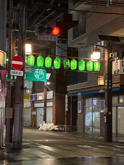 This media file depicts a nighttime scene in what appears to be a narrow street or alleyway in Adachi, Japan. The image is dominated by a series of bright green lanterns hanging vertically from overhead wires, illuminating the otherwise dim surroundings. To the left, a traffic light is clearly visible, showing a red signal, indicating that vehicles should stop. Below the traffic light, a sign reads "自転車専用" (Jitensha Sen'yō), meaning "Bicycle Exclusive" or "Bicycle Lane". Further down, another sign says "自転車を除く" (Jitensha o Nozoku), which translates to "Excluding Bicycles".
The architecture of the buildings lining the street is visible, with various shop fronts and windows. Some signs with Japanese text are present, including "千住宿" (Senjuku), which might refer to a location or a historical district. The ground appears wet, suggesting it may have rained recently or the street is being cleaned, reflecting the ambient light. There are no people clearly visible in the frame, creating a sense of quietness and solitude. The overall atmosphere is characteristic of a Japanese urban environment at night, with a focus on the illuminated lanterns and signage. The presence of "UR" and "BUNGU KAMIYA" on some banners suggest commercial establishments. The scene is likely a street that caters to local residents or visitors, with clear regulations for bicycle traffic.