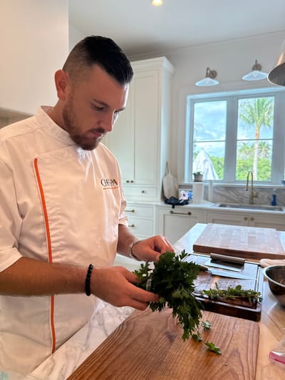 In East Naples, Florida, a Caucasian male chef, dressed in a white chef's coat with orange trim and a logo that reads "CHEPA," is preparing herbs in a well-lit kitchen. He has dark hair styled in an undercut and a beard, and is wearing a beaded black bracelet on his left wrist. He is holding a bunch of fresh parsley in his hands and appears to be separating the leaves. In front of him, on a marble countertop, are several wooden cutting boards, a knife, and a metal bowl. Behind the chef, a large window offers a view of lush green palm trees and a blue sky. The kitchen is equipped with white cabinetry and a gold-toned faucet.