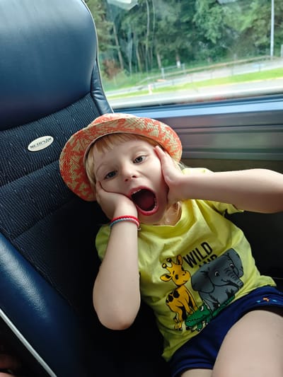 A young child with blonde hair, wearing a patterned hat and a yellow t-shirt with animal graphics, sits in a dark blue bus seat. The child has their hands on their cheeks and their mouth is wide open, as if surprised or shouting. A bracelet with red and blue beads is visible on their left wrist. In the background, through a window, trees and a road are visible, suggesting the bus is in motion, possibly on a street in Eugendorf, Austria, given the context of the image's origin. The "NEOPLAN" logo is visible on the headrest of the seat.