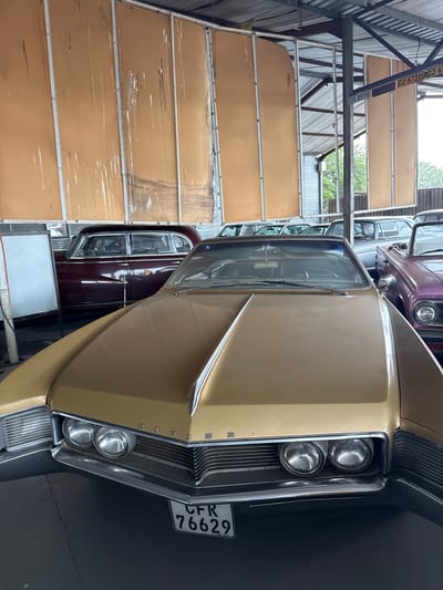 The image shows a close-up of the front of a vintage gold-colored car, likely a Buick Riviera, parked indoors. The car has a chrome grille with four headlights on each side and a license plate reading "CFR 76629." Behind the gold car, another vintage car is visible, this one maroon. The location is in the city of Kraaifontein, South Africa, based on license plate. A wall structure made of metal frames with panels can be seen in the background, suggesting a garage or storage facility. No people are visible in the image.