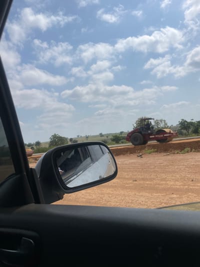 The image shows a view from inside a vehicle, with a side mirror reflecting the surroundings. Outside the vehicle, there is a road roller compacting a dirt road in Dokwa, Nigeria. The sky is visible with scattered clouds. The landscape appears arid, with sparse vegetation. The interior of the car includes the door panel and window frame.