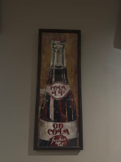 A horizontally oriented framed print shows a stylized depiction of a vintage Coca-Cola bottle. The bottle is rendered in shades of red, white, and black, with aged or distressed effects. The iconic “Cola Up” label is prominently featured. The background is a mottled golden-brown, suggesting a vintage or rustic aesthetic. The frame is dark and rectangular. The artwork is mounted on a neutral-colored wall.