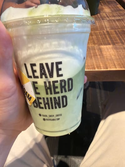 The image shows a hand holding a cup of a beverage. The cup appears to be clear plastic with a clear lid and contains a light green and white layered drink. The label on the cup reads "LEAVE THE HERD BEHIND," with additional text indicating "BLACK SHEEP COFFEE" and "RECYCLABLE CUP." A sticker reading "FOAM" is also visible. The background includes a wooden surface and a draped white fabric. The environment suggests an indoor setting. There is no mention of the location of Bretton, United Kingdom, in the image.