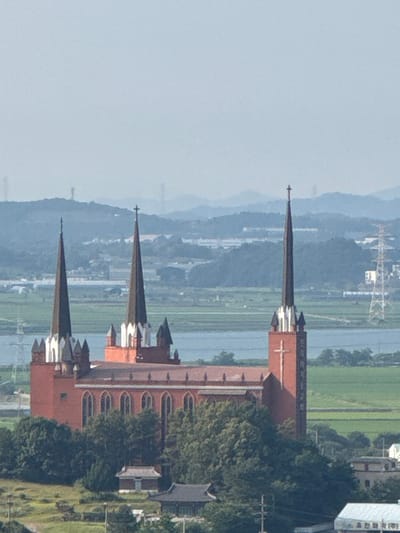 A high-angle, full shot captures a large, red brick church with multiple spires, characteristic of Gothic architecture, set against a backdrop of rolling hills and a hazy sky in Pyeongtaek-si, South Korea. The church features tall, pointed spires with crosses at their apexes and arched windows along its facade. The building is surrounded by lush green trees and vegetation, with a body of water, possibly a river or lake, visible in the middle ground. In the distance, power lines and other structures hint at a populated area. The overall scene depicts a serene and majestic architectural landmark within a natural landscape.