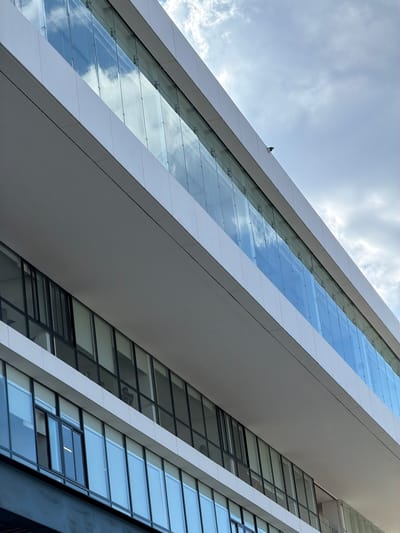 A modern, multi-story building facade in Fraccionamiento Los Heroes Queretaro, Mexico, is depicted under a partially cloudy sky. The structure exhibits clean lines with prominent white horizontal fascias and extensive glazing. The uppermost visible level features a continuous glass railing, secured by metal standoffs, reflecting a blue sky with white, scattered clouds. A dark, dome-shaped security camera is mounted on the building's top white fascia, positioned centrally above the upper glass railing. Below a substantial white horizontal overhang, a lower floor is visible, comprising numerous rectangular windows with dark frames. These lower windows display varying transparency, with some reflecting the sky and others revealing dimly lit interior spaces. A horizontal light source is visible within a window pane on the lower left. Vague dark forms, consistent with human figures, are discernible behind several of the lower windows.