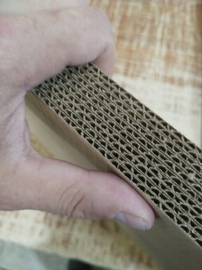 The image shows a close-up of a person's hand holding a piece of corrugated cardboard. The cardboard is layered with a wavy, corrugated inner material sandwiched between two flat outer surfaces. The hand appears to be gently pressing on the cardboard, with the fingers and thumb visible. The background is blurred, but appears to be a wooden surface. There is no indication of a specific location in the city of Гожскі сельскі Савет, Belarus. The scene is simple and focuses on the texture and material of the cardboard and the human interaction with it.