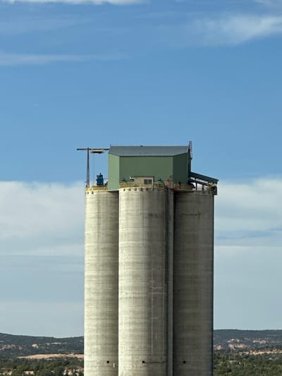 The image depicts three cylindrical concrete grain silos that are part of a industrial facility. The silos are tall and stand against a blue sky with scattered clouds. A green building is situated on top of the silos, connecting them. A metal structure with a crane-like component extends from the left side of the green building, over one of the silos. The facility is located in an arid, hilly landscape with sparse vegetation. The image appears to have been taken from a distance, focusing on the silos and their upper structure. There are no people visible in the image. The scene is likely located in Shonto, United States, based on the visual context of arid terrain and industrial structures.