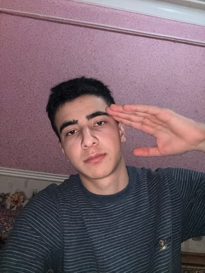 A young man with dark hair is featured in the image. He wears a striped, dark-colored shirt. His right hand is raised, palm facing forward, positioned near his forehead in a gesture resembling a salute. The background consists of a light purple or pink wall. The man looks directly at the camera. The image appears to be rotated, as the wall is on the left and top of the frame, and a glimpse of flooring is visible on the right.