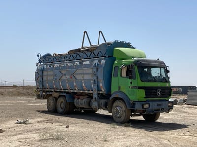 A green and blue semi-truck with a large tank on the back is parked on a dirt lot in Pir Dust, Afghanistan. The truck is facing to the right and is angled slightly. The truck's wheels are all on the ground. There is a blue and white sign on the front of the truck. Behind the truck is a pile of gray metal objects. The truck is sitting in a large open lot, with a clear blue sky above. The ground is covered in light-colored sand. There are no people visible in the photo. The location is an open dirt lot within Pir Dust, Afghanistan, specifically at coordinates (latitude: 31.633, longitude: 65.854).