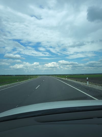 The image captures a view from a vehicle driving on a paved road. The road curves to the right, with white dashed lines marking the lanes. On the left side of the road, there is a field of green crops, and on the right, a field of dark soil. White and red striped posts line the shoulder of the road. The sky is filled with fluffy white clouds against a blue background. The foreground shows the dashboard and windshield of the vehicle. The scene appears to be in a rural area, possibly near Zol'skaya, Russia, given the agricultural landscape and the presence of road markers typical of Russian roads. There are no people visible in the image.