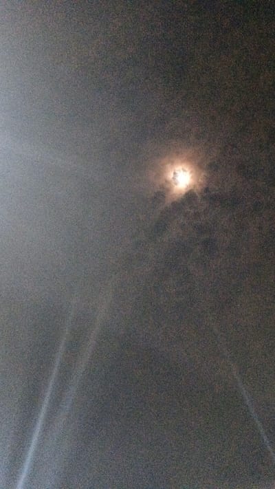 The image shows a hazy, nighttime sky with the moon partially obscured by clouds. The moon appears as a bright, luminous orb with wisps of dark clouds swirling around it. The sky is dark gray and filled with digital noise. In the lower left and right corners, two bright, diagonal shafts of light pierce through the darkness, suggesting a strong light source from below the frame. The overall scene is atmospheric and somewhat mysterious, with the light beams adding a dramatic element to the cloudy night sky. The specific location of Uteungeulinggang, Indonesia, is not visually represented in the image.