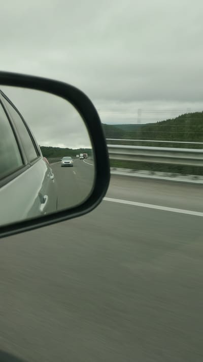 The image is a side-view mirror reflection of a highway in Baliovtsi, Bulgaria. In the reflection, a silver car is visible in the distance, followed by a red truck. The highway has white lane markings and a metal guardrail on the right side. Beyond the highway, there are green trees and a cloudy sky. The overall scene suggests a drive on a cloudy day.