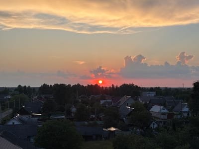 The image captures a sunset over a rural village, likely in the Protasevichi rural council of Belarus. The sky is a gradient of warm colors, transitioning from orange and yellow at the top to pink and red near the horizon. Several clouds are scattered across the sky, some illuminated by the setting sun, creating dramatic silhouettes. The sun itself is a bright, fiery orb partially obscured by a cloud, casting a warm glow over the scene.
Below the sky lies a dense canopy of trees that partially hide a collection of houses. The houses appear to be typical rural dwellings with dark, sloped roofs, some with visible chimneys. Some houses have visible windows, and a few have illuminated lights, suggesting that it is either early evening or the houses have internal lighting. A road can be seen on the left side of the image, snaking through the village and disappearing into the trees. The overall atmosphere is serene and peaceful, with the vibrant colors of the sunset contrasting with the subdued tones of the village. There are no people or animals visible in the frame. The image is taken from an elevated viewpoint, offering a panoramic view of the village and its surroundings.