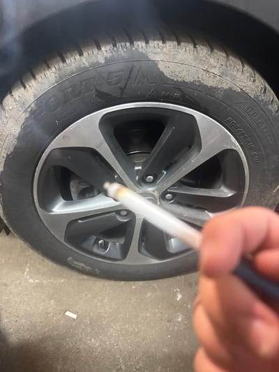 The scene is situated in Maslarevo, Bulgaria. The image presents a close-up, low-angle perspective focusing on a lit cigarette held by a human hand in the foreground, with a vehicle's front passenger side wheel as the primary background subject.
The ground appears to be a concrete or asphalt surface, scattered with light dust or debris. A distinct white cigarette filter or butt lies on this surface beneath the car wheel, towards the lower-left.
A right human hand, with fingers partially visible, holds a lit cigarette diagonally across the mid-frame. The cigarette's tip is amber, indicating it is actively burning, and a thin stream of white smoke emanates from it, rising towards the upper-left where it dissipates into a larger, hazy plume.
The car wheel consists of a black rubber tire and a five-spoke alloy rim. The tire, identified as "SPORT 5 M+S," displays markings including "RADIAL TUBELESS," a "ROTATION" arrow, and size specifications "205/55R16 91H." Multiple white snowflake symbols are visible on the tire's sidewall, indicating its winter classification. The tire's surface, particularly the tread and portions of the sidewall, is coated with dried mud or dirt. The alloy rim features a two-tone design with dark grey/anthracite inner spokes and silver/machined outer edges, secured by five silver lug nuts. A brake disc is partially visible behind the rim spokes.