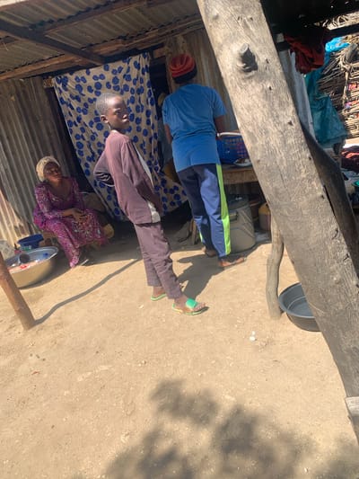 In Project, Nigeria, a scene unfolds under a makeshift, corrugated metal roof supported by rough-hewn wooden beams. The ground is a mixture of compacted dirt and sand. In the mid-ground, a young person with short dark hair, wearing a dark brown long-sleeved shirt and dark pants, stands facing left, looking towards the camera. They are wearing light green flip-flops. To their left, a woman in a vibrant purple, intricately patterned dress and a light-colored headscarf sits on a low wooden bench. Before her, a large metal basin contains water and a bottle. Behind the young person, another individual, partially obscured, wears a bright blue t-shirt and dark pants with a green stripe down the side, along with a red beanie. They are looking towards a blue plastic basket on a wooden table. To the far right, a thick, weathered wooden post dominates the foreground, casting a strong shadow. On the ground near this post is a dark basin filled with water. The background reveals more of the rustic shelter with draped fabric and various items, suggesting a functional, outdoor living or working space.
