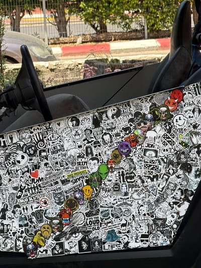 The image depicts the dashboard of a vehicle covered extensively with a variety of black and white stickers. There are also a variety of colorful buttons placed around the dashboard. The vehicle is likely located in Kiryat Motzkin, Israel based on the setting visible through the windshield, which includes a sidewalk with a red and white painted curb, a metal fence, and foliage. A portion of the vehicle's steering wheel and seat are visible.