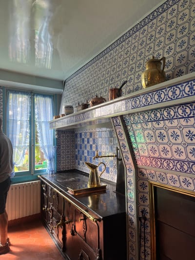 The image shows the kitchen of Claude Monet's house in Giverny, France, located at the coordinates 49.075, 1.534. The room has a large, old-fashioned black stove with several ovens. The stove is positioned along a wall decorated with white and blue ceramic tiles, a common decorative element in French kitchens. The backsplash above the stove features an elaborate, tile-covered mantle with a copper kettle, a brass pitcher, and other assorted metal cookware. The kitchen appears to be unoccupied, with no people present. A window with blue gingham curtains is visible to the left of the stove, which shows a glimpse of the exterior garden. The ceiling features a glossy white paint with some faint, light-colored reflections.