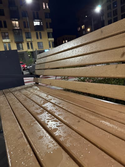 An exterior shot at night, focusing on a wet wooden park bench in Laikovo, Russia. The bench's slats are covered in numerous water droplets, indicating recent rain. In the background, a modern residential building with lit windows stands under a dark sky. A street light illuminates the scene. Partially visible in the mid-ground is a dark structure to the left, and beyond that, two cars are parked, one red and one gray. Green foliage is present behind the bench. The perspective is low, emphasizing the bench's texture and the reflections of light on the wet wood.