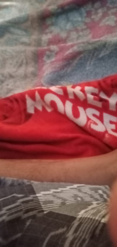The image shows a section of a patterned bed covering in the city of Bulukandang, Indonesia. Over the bed covering is a red t-shirt with white lettering that reads "Mickey Mouse". Adjacent to the shirt is a person's arm, which is partially out of focus.