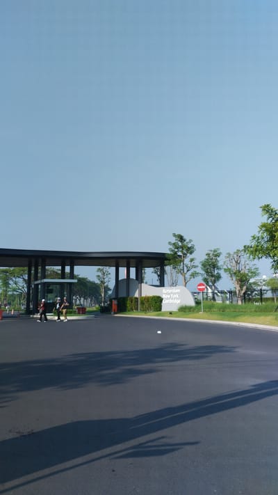 The image depicts the entrance to a residential area in Trosobo, Indonesia, identified by a sign that reads "Rotterdam New York Cambridge." The entrance features a modern, open-air structure with a dark roof supported by pillars. In front of this structure, a security booth is visible. Three individuals, appearing to be security personnel, are standing near the entrance, dressed in uniforms and hats.
To the right of the entrance structure, a curved, light-colored monument or sign is positioned. This monument is adorned with lush green bushes and features the aforementioned text. Adjacent to the monument is a red and white circular traffic sign.
The foreground of the image is dominated by a paved road, reflecting the shadows of trees, suggesting it is a sunny day. The road curves to the right, leading away from the entrance. Beyond the entrance, rows of trees and a black fence line the perimeter of the property. The sky is clear and blue, occupying the upper half of the frame.