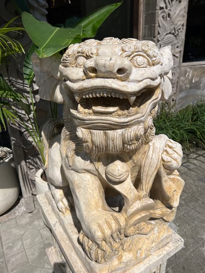 A pale stone statue of a mythical beast with an open mouth and sharp teeth stands outdoors. The sculpture is intricately detailed, with textured fur-like patterns. The beast's nose has prominent holes. Foliage flanks the statue on the left. The background includes a building with carved decorative elements and surrounding greenery. The statue rests on a stone base. The setting appears to be the city of Petulu, Indonesia.