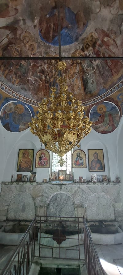 The image depicts the interior of a building, possibly a church or monastery, in Бачково, Bulgaria. The view is angled upwards. The ceiling is adorned with frescoes and paintings, including circular depictions of figures. A large, ornate gold chandelier hangs in the center of the frame. The right side of the image shows stone walls with a series of religious icons mounted on the wall. A metal railing is visible, suggesting a balcony or upper level. The scene is dimly lit with light filtering through a window.