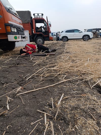 An outdoor scene in Kottalapalli, India, depicts a male individual lying prone and motionless on dark, cracked earth intermingled with dry, light-colored plant stalks. The individual is dressed in a red uniform jacket with striped cuffs and collar, dark trousers, and a dark cap. A long, dark-colored rifle with a wooden stock lies on the ground parallel to the individual's left side. In the background, several vehicles are parked on the edge of the dry field: an orange truck, partially visible on the far left, bears the text "GENERATOR POWERED" on its bumper; a second orange truck with an open cab door and a tarp-covered bed is positioned behind it. Further to the right, a white SUV is parked, and another light-colored vehicle is partially visible in the far background. The sky is overcast.