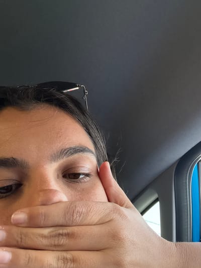 A close-up image captures the upper portion of a person's face inside a vehicle, located in West Windsor, United States. The subject has dark hair and brown eyes, with dark-rimmed sunglasses perched on top of their head. Their right hand is positioned across the lower part of their face, covering the mouth and nose area, with the thumb resting near the right cheek. The interior of the vehicle is visible in the background, featuring a dark gray ceiling and a car window frame on the right, through which a light blue exterior element and a bright, possibly outdoor, scene are partially discernable.