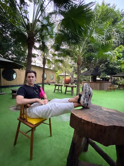 A person sits in a chair on a green surface. They are wearing a black shirt, white pants, and sneakers. Their feet are resting on a wooden table. The chair has a yellow cushion. Behind the person is a building with round windows. In the background, there are trees, some of which are palm trees. The image was taken in Nam Nha Trang, Vietnam.