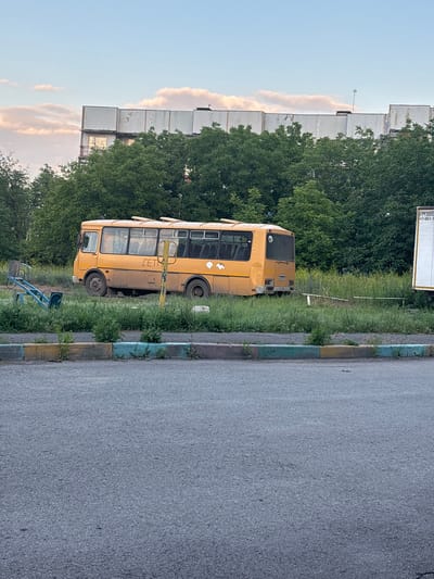 An abandoned yellow bus, likely a former school bus given the Cyrillic lettering "ДЕТ" (likely part of "ДЕТСКИЙ" meaning "children's") visible on its side, sits in a grassy field next to a blue seesaw playground structure. The bus is dusty and appears weathered, with some graffiti or stickers on its rear. In the background, a large, unfinished concrete building with scattered trees in front of it rises against a sky with scattered clouds. A white truck with some Cyrillic text, possibly a freight or advertisement truck, is partially visible to the right of the bus. The foreground shows a paved road and a low curb painted with alternating yellow, green, and blue sections. The scene suggests an area on the outskirts of Gukovo, Russia, possibly a disused public space or the grounds of an abandoned facility. There are no people visible in the image.