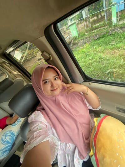 Inside a vehicle with an open sunroof, a young woman wearing a pink hijab and floral dress leans back against a car seat. She is smiling and holding up two fingers in a peace sign. Outside the sunroof, trees and buildings are visible. A large, plush hamburger toy sits beside her. Her feet are visible near a blue pillow and a red shoe. The angle suggests the photo was taken from above.