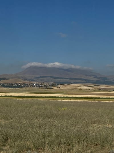 The image displays a panoramic view of the landscape surrounding Ortaköy, Turkey. In the foreground, a field of tall, dry grass stretches across the frame. Beyond this field, a pattern of agricultural land unfolds, with distinct sections appearing as fields of varying colors, suggesting different crops or stages of growth. A thin, pale yellow strip runs horizontally across the middle ground, possibly a road or a different type of field.

Dominating the background is a large, imposing mountain. The summit of the mountain is partially obscured by a band of white, fluffy clouds that hug its peak. Nestled at the base of the mountain, a small village is visible. The cluster of buildings, characterized by red-tiled roofs and white walls, is interspersed with green trees. A slender white minaret, a common feature of mosques in Turkey, rises above the village buildings, a prominent landmark. The sky above is a clear, vibrant blue, with a few wispy clouds scattered in the upper portion. The overall impression is one of a rural, mountainous region in Turkey.