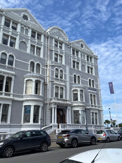 The image shows the exterior of the Imperial Hotel in Llandudno, United Kingdom. The hotel is a large, gray building with white architectural details, including arched windows, decorative molding, and columns. A flag is visible on the roof of the building. Cars are parked along the street in front of the hotel. The sky is blue with scattered clouds.