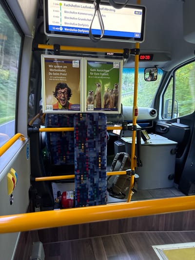 The image depicts the interior of a bus in Schuld, Germany. A digital display above shows bus route information and the current time, 09:37. Below, two advertisements are visible. The left ad features a man with glasses and the text "Wir sparen am Werbemodell, Du beim Preis!" and "25% auf Einzeltickets sind die beste Werbung.". The right ad shows meerkats and the text "Gute Aussichten für Eure Freizeit." and "Bis zu 5 sind günstig unterwegs.". A digital clock on the bus ceiling shows the time as 09:38. The bus seats are upholstered with a blue, red, and yellow patterned fabric. A red fire extinguisher is visible on the floor. Outside the bus window, a blurred green landscape can be seen.