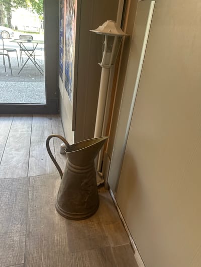 The image depicts the interior of a establishment in Molenbeek-Saint-Jean - Sint-Jans-Molenbeek, Belgium, near a doorway that opens to an outdoor seating area. On the interior floor, which is tiled with a wood-like pattern, a decorative metal pitcher with a handle sits to the left. Behind the pitcher, a tall, white outdoor lamp post stands vertically. The wall to the right of the lamp post is a light beige color. Through the doorway, an outdoor scene is visible with a parked car on a street, a small table with chairs, and trees. The overall impression is of a cafe or restaurant entrance area.