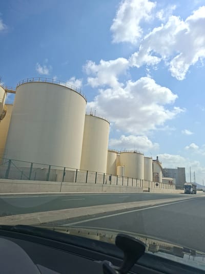 This is a landscape, eye-level, medium shot photograph taken from a moving vehicle. The scene depicts a row of large, cylindrical storage tanks, likely industrial silos, with a light yellow or cream exterior. They are positioned behind a concrete barrier topped with a metal fence. The sky above is a bright blue with scattered, fluffy white clouds, suggesting fair weather. A roadway is visible in the foreground, with lane markings and a concrete divider. In the distance, a white truck is driving on the road, and industrial buildings and structures are present. The overall impression is of an industrial area on the outskirts of a city. This industrial complex is located in the Escombreras Valley, a significant industrial zone near Cartagena in Murcia, Spain, known for its refineries and chemical plants.